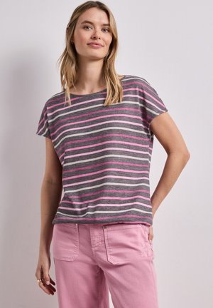 Frau trägt ein grau-, rosa- und weiß gestreiftes kurzärmeliges Oberteil und hellrosa Hose, steht mit einer Hand in der Tasche an einer schlichten Wand.