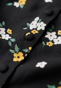 Tissu noir avec un petit imprimé floral jaune et blanc et des boutons noirs recouverts de tissu le long d’une couture.