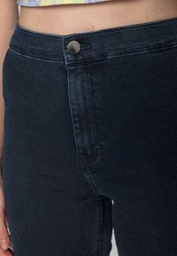 Mörkblå denimjeans med en figurnära passform, som har en framficka, knappstängning och subtil textur. Närbild på midjebandet.