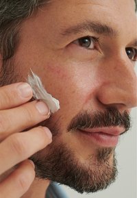 Facial moisturizer applied to the cheek of a man with a well-groomed beard. Skin appears smooth with slight redness, light color cream used.