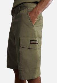 Shorts cargo vert olive en tissu léger, avec des poches latérales et un patch de marque visible sur la cuisse gauche.
