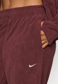Burgundské flísové tepláky s elastickým pásom a bočnými vreckami, s nenápadným bielym logom Nike na ľavom stehne.