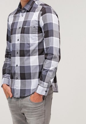 Mann trägt ein schwarz-grau-weiß kariertes Hemd mit Knöpfen und hat die Hände in den Taschen einer hellgrauen Jeans vor einfarbigem Hintergrund.