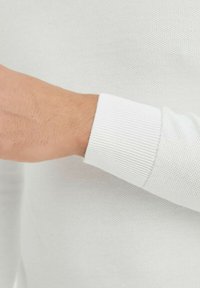 Vêtement tricoté à manches longues de couleur blanche, vu du poignet vers le haut, avec un poignet côtelé et une surface texturée en tissu léger et respirant.