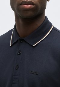 Άνδρας που φοράει μπλε ναυτικό πόλο μπλουζάκι με λευκό και μπεζ ριγέ γιακά και τρία κουμπιά, με ανάγλυφο λογότυπο «BOSS» στο στήθος.