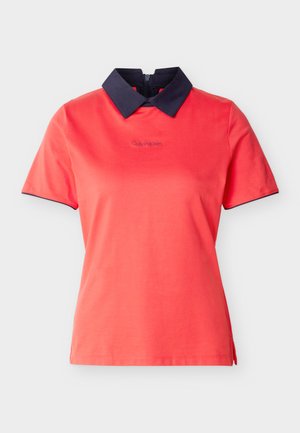 Korálově červená košile s krátkým rukávem, černým špičatým límečkem, malým logem Calvin Klein na hrudi a černým lemováním na okrajích rukávů.