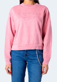 Femme portant un sweat-shirt rose Levi's et un jean bleu avec une chaîne argentée pendant de la poche, sur un fond uni.