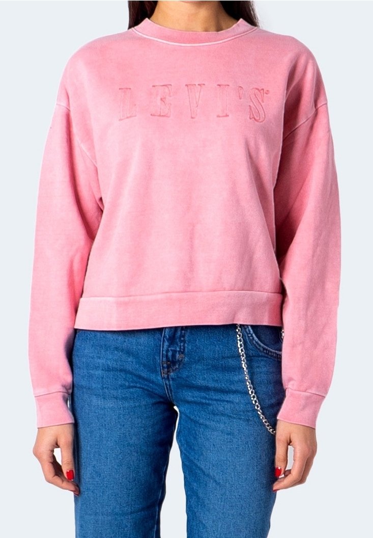 Femme portant un sweat-shirt rose Levi's et un jean bleu avec une chaîne argentée pendant de la poche, sur un fond uni.