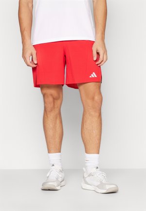Mężczyzna w czerwonych sportowych szortach z logo Adidas, białych skarpetkach i białych butach sportowych, stojący na jednolitym tle.