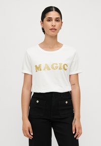 White short-sleeve t-shirt featuring the word "MAGIC" in bold, shiny gold lettering. Black high-waisted trousers with front pockets.