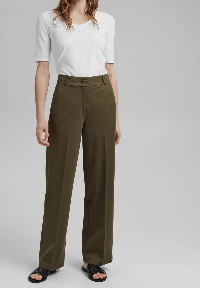 Femme portant un pantalon large vert olive, un top blanc à manches courtes côtelé, et des sandales noires ouvertes à l'avant, debout devant un fond gris.