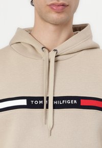Bežinė kapučino su raišteliais, su horizontalia mėlyna juostele, puošta raudonomis ir baltomis detalėmis. „TOMMY HILFIGER“ tekstas baltai ant mėlynos juostelės.