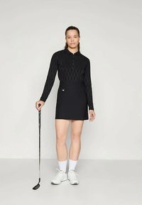 Mladá žena stojí s golfovým putterom, oblečená v čiernom tričku s dlhým rukávom, čiernej sukni, bielych ponožkách a bielych golfových topánkach na jednoduchom pozadí.