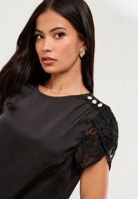 Čierna blúzka s krátkymi rukávmi a čipkovými detailmi na pleciach, hladká látka, tri dekoratívne gombíky na pleci a okrúhly výstrih.