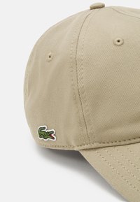 Khaki bejzbol cap s zakrivljenim vizmi, napravljen od teksturirane tkanine. Ima zeleni zakrpa s logotipom krokodila sa strane.