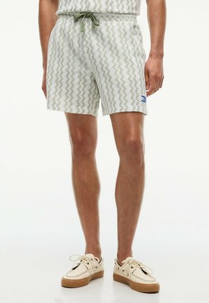 Mann trägt helle gemusterte Shorts mit grünem Kordelzug und beigen Freizeitschuhen mit Gummisohlen, steht vor einem einfarbigen Hintergrund.