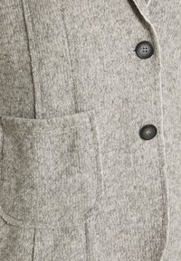Chaquetón de punto gris claro con cuello de muesca, dos botones y un bolsillo frontal con bordes sin acabar y una textura sutil.