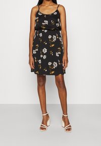 Czarna sukienka w kwiaty z cienkimi ramiączkami, zdobiona białymi i żółtymi kwiatami. Luźny krój z elastyczną talią, zestawiona z białymi sandałami na słupku.