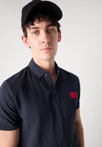 Mørkeblå polo skjorte med tre knapper og ribbestrikket krage. Har et rødt rektangulært logo-patch merket "HUGO" på venstre bryst.