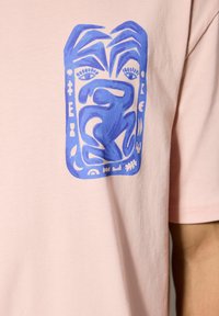Camiseta de algodón rosa claro con un diseño gráfico azul que presenta formas y símbolos abstractos, ubicado en la zona del pecho.