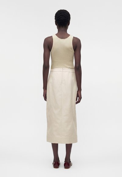 Άτομο που φοράει μπεζ αμάνικη ριμπ μπλούζα, κρεμ μίντι φούστα και σκούρα κόκκινα παπούτσια, στέκεται γυρισμένο με την πλάτη σε ένα απλό φωτεινό φόντο.