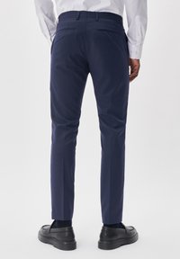 Marinblå skräddarsydda byxor, smal passform, med en slät textur, bälteshällor och två bakfickor. Matchas med svarta slip-in skor.