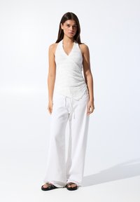 Top in tessuto bianco a halter con effetto testurizzato, abbinato a pantaloni bianchi larghi e sandali neri con dettagli decorativi.
