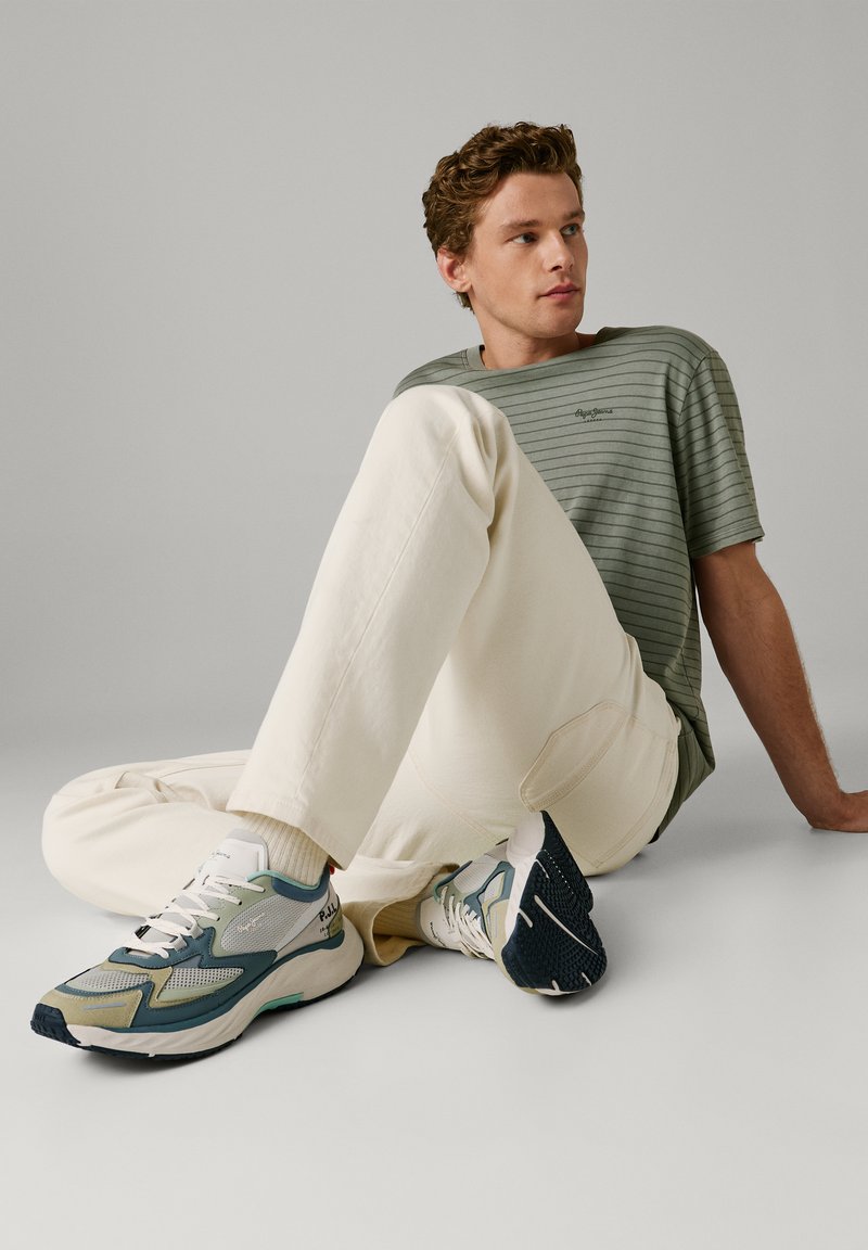 Jeune homme assis par terre portant un t-shirt à rayures vertes, un pantalon crème et des baskets grises et bleues, regardant légèrement sur le côté.