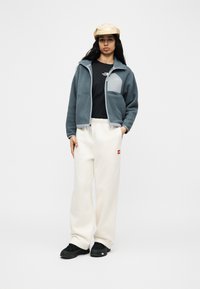 Γυναίκα που φοράει μπεζ καπέλο, μπλε fleece με φερμουάρ, μαύρο μπλουζάκι, λευκό φαρδύ παντελόνι και μαύρα παπούτσια, στέκεται με το ένα χέρι στην τσέπη.