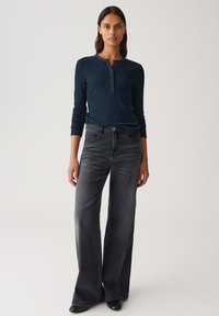 Frau trägt ein navyfarbenes, geripptes Langarm-Henley-Shirt und hochtaillierte, ausgewaschene schwarze Weitleggings, steht vor einem schlichten Hintergrund.