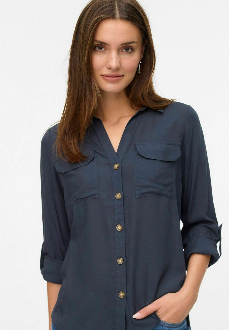 Chemise boutonnée à manches longues bleu marine, avec deux poches poitrine, manches retroussées et boutons dorés. Tissu doux et lisse.