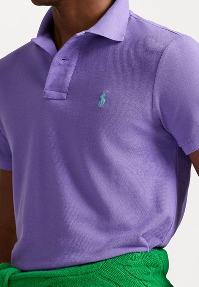 Purpurna polo majica iz teksturiranega blaga, z turkiznim logotipom na levem prsnem delu; v kombinaciji z zelenim pletenim pasom.