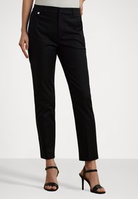 Zwarte op maat gemaakte broek van soepel materiaal, met een slim fit en plooidetail. Gecombineerd met zwarte hoge hakken en een lichte top.