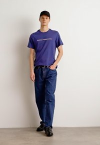 Navy blue t-shirt with "BARBOUR INTERNATIONAL" logo, paired with loose blue jeans and black shoes. Person wears a black cap.