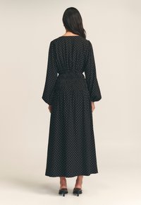 Black long-sleeve dress with white polka dots, gathered waist, and a flowing silhouette. Smooth fabric with a relaxed fit.
