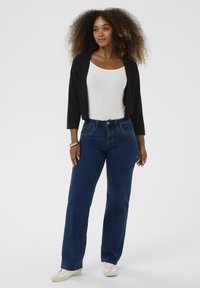 Eine Frau steht in einem schwarzen offenen Cardigan, einem weißen Tanktop, dunklen blauen Jeans, weißen Sneakers und einem silbernen Armband an ihrem linken Handgelenk.