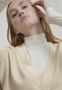 Top beige con un design a V sovrapposto a un leggero dolcevita, che mette in mostra una texture di tessuto liscia e maniche delicatamente drappeggiate.