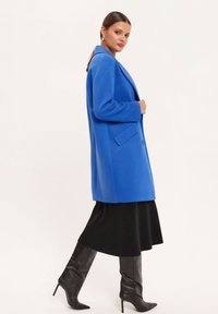 Kobieta w jasnoniebieskim płaszczu, nosząca czarną sukienkę i czarne wysokie kozaki za kolano, stoi w profilu na białym tle.