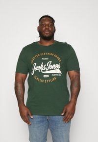 Mørk grønn bomulls T-skjorte med rund hals, med hvit og oransje grafisk tekst: "Jack & Jones" og "Klassisk stil." Korte ermer.