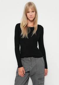 Tricou negru cu mânecă lungă, cu decolteu tip scoop, asociat cu pantaloni cargo gri. Materialul pare moale și are o siluetă ajustată.