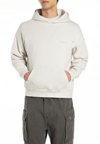 Sudadera con capucha de color beige claro en una tela suave y texturizada, con un bolsillo frontal y puños acanalados. Combinada con pantalones cargo grises.