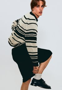 Maglione a maglia con strisce nere e crema, abbinato a pantaloni corti neri. Il modello indossa scarpe nere e calzini grigi chiari, seduto con una gamba sollevata.