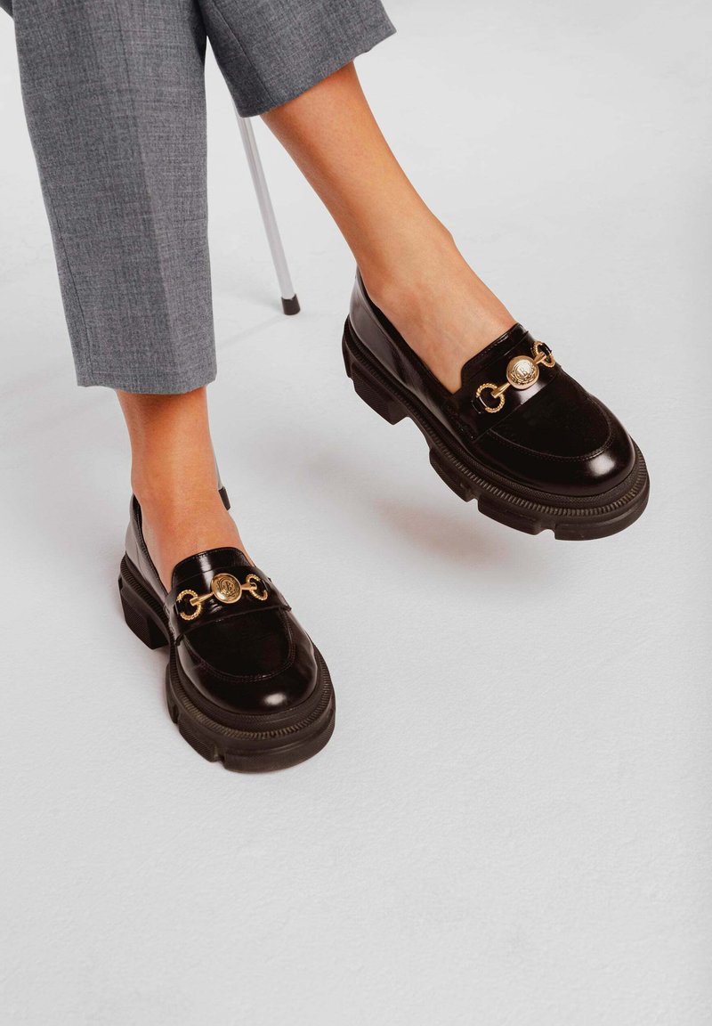 Zwarte lakschoenen met een stevige zool, gouden kettingaccent en ronde metalen details aan de voorkant. Getextureerde stoffen broek zichtbaar.