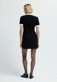 Donna con capelli biondo corto che indossa un vestito nero a maniche corte con colletto e polsini bianchi, collant neri trasparenti e scarpe nere, in piedi.