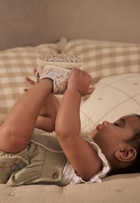 Scarpine in pelle con stampa floreale, colori chiari e cinturino regolabile. Braccia di un bambino sollevate, sdraiato su biancheria da letto beige a quadri.