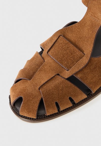 Sandal en suède marron avec un design découpé, bout ouvert et accents marron foncé, dotée d'une semelle texturée. Sangle ajustable pour un maintien sécurisé.