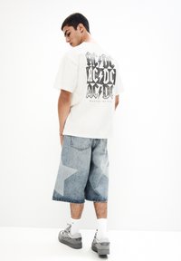 Bílý tričko s oversized grafikou v černé barvě, se nápisem "AC/DC." Na sobě má světle modré džínové kraťasy s vzorem světlejších hvězdiček a šedé tenisky.