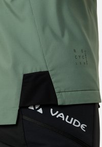 Giacca da ciclismo verde con una superficie texture, caratterizzata da un pannello nero nella parte inferiore e un logo bianco. Include dettagli di branding per il riciclo.