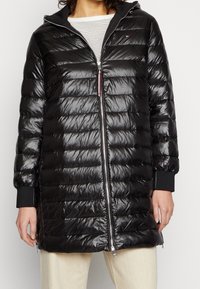 Czarna kurtka puchowa o błyszczącym wykończeniu, zapinana z przodu na zamek, z kapturem, ściągaczami przy mankietach oraz poziomym pikowaniem. Pod spodem widoczna jest w paski koszula.