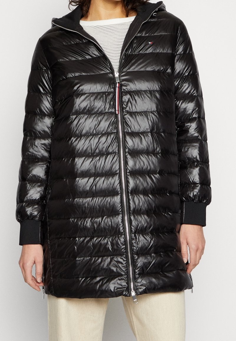 Czarna kurtka puchowa o błyszczącym wykończeniu, zapinana z przodu na zamek, z kapturem, ściągaczami przy mankietach oraz poziomym pikowaniem. Pod spodem widoczna jest w paski koszula.